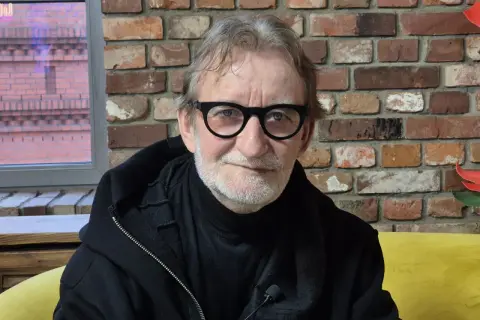 Maciej Mańkowski w okularach, z siwą brodą, w czarnej bluzie na tle ceglanej ściany.
