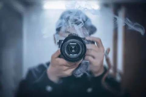 Osoba w kapturze śledzi i fotografuje gwiazdy przez obiektyw aparatu, otoczona mgiełką dymu.