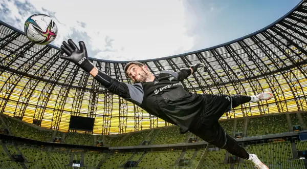 Bramkarz w locie łapie piłkę. Dynamiczna fotografia sportowa na tle stadionu.