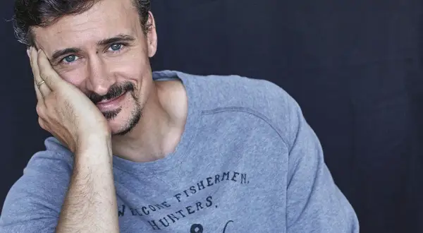 Mateusz Dela zamyślony, opiera głowę na dłoni. Na szarym swetrze widnieje napis "WELCOME FISHERMEN, HUNTERS".