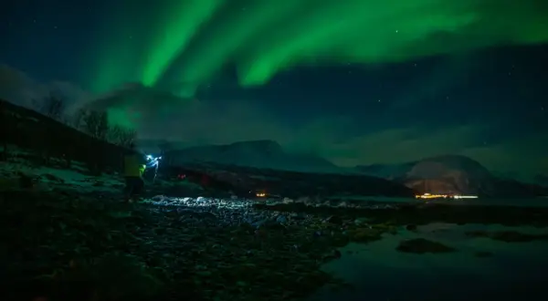 Nocne niebo rozświetla zorza polarna. Na pierwszym planie widać fotografa z lampą, który robi zdjecie z fleszem.