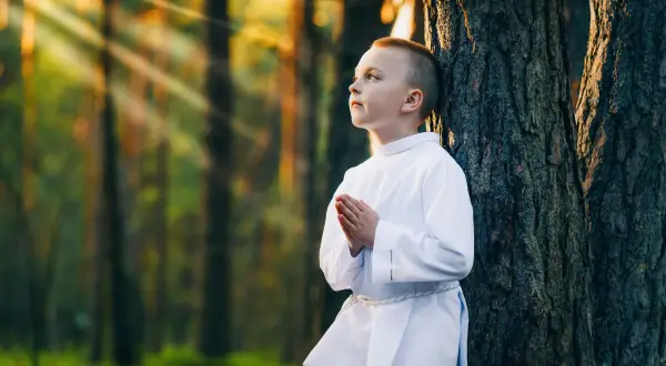 Chłopiec w bieli oparty o drzewo, modlący się. Promienie słońca przebijają się przez las. Piękne sesje komunijne w plenerze.