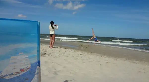 Dziewczyna robi zdjęcie akrobatce na plaży. W tle widać morze i chmury, a horyzont jest lekko krzywy.