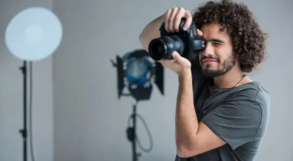 Mężczyzna z kręconymi włosami i aparatem w dłoniach, ćwiczący, jak zostać fotografem. W tle lampy studyjne.