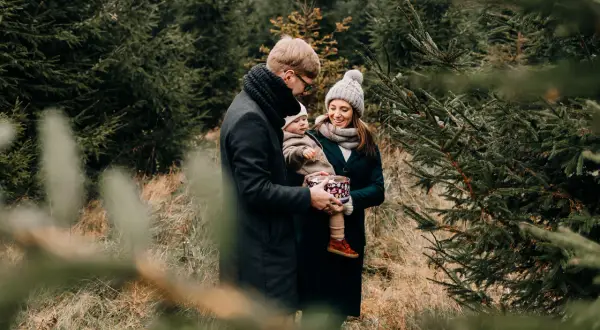 Rodzina podczas sesji świątecznej w plenerze. Ojciec trzyma dziecko, matka uśmiecha się, otoczeni choinkami.