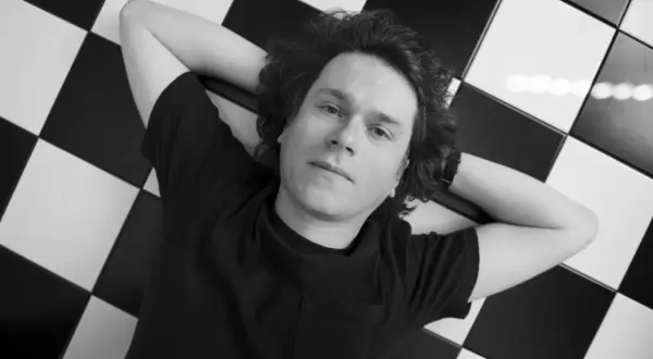 Hubert Humka leży na czarno-białej szachownicy, ręce za głową, w czarnej koszulce.