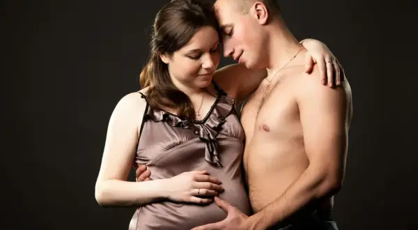 Czuła jesienna sesja ciążowa. Para obejmuje się czule, mężczyzna delikatnie dotyka brzucha kobiety.