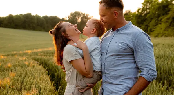 Szczęśliwa rodzina podczas sesji na łące. Mama śmieje się, trzymając dziecko, a tata patrzy na nich z czułością.