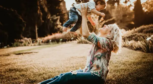 Mama rzuca dzieckiem w górę podczas śmiesznej sesji rodzinnej w parku. Słońce zachodzi, tworząc ciepłą, złotą poświatę.