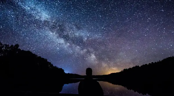 Sylwetka człowieka patrzy na Drogę Mleczną nad jeziorem. Idealny kadr, by pokazać, jak fotografować gwiazdy.