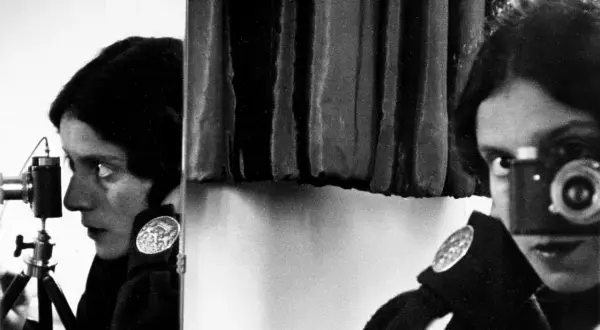 Tina Modotti w kadrze, z aparatem w ręku, patrzy w obiektyw.