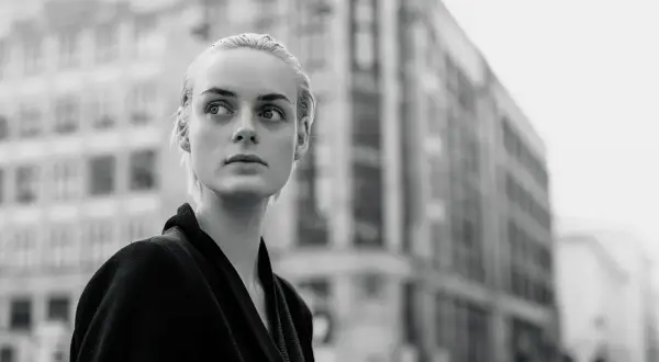 Monochromatyczna fotografia. Młoda kobieta o blond włosach patrzy w bok, na tle miejskiej architektury.