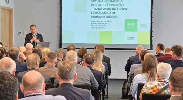 Mężczyzna przemawia na spotkaniu roboczym. Na ekranie widnieje napis "Spójna promocja polskiej żywności". Może to być wada obiektywu, która lekko zniekształca obraz.