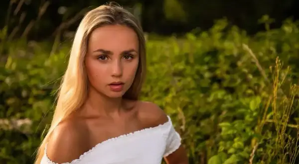 Młoda kobieta z długimi blond włosami, ubrana w białą, odkrywającą ramiona bluzkę, patrzy prosto w obiektyw na tle zielonej, rozmytej roślinności.