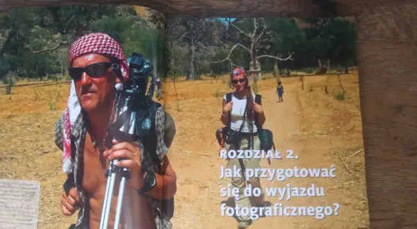 Piotr Trybalski z aparatem i statywem w gorącej Afryce. Obok niego kobieta i dziecko na pustynnej drodze.