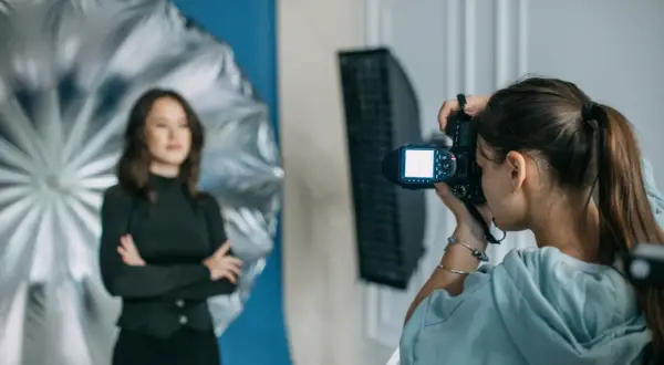 Dzień fotografa w studiu. Młoda kobieta z aparatem w ręku, w tle modelka pozująca na tle niebieskiego tła i srebrnego parasola.