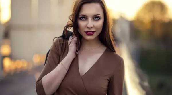 Piękna kobieta w brązowej sukience, z włosami opadającymi na ramię. Jej spojrzenie przyciąga uwagę w tej subtelnej fotografii portretowej.