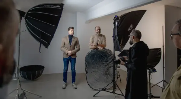 W studiu fotograficznym trwa sesja. Widać ludzi, sprzęt i modyfikatory światła w fotografii, w tym parasole i blendę.