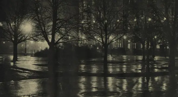 Nocny park po deszczu, drzewa bez liści, mokre ścieżki odbijające światła budynków. Alfred Stieglitz uchwycił atmosferę miasta.
