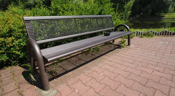 Szeroki kadr ukazuje pustą, metalową ławkę w parku, obok której leży aparat fotograficzny. W tle zielone drzewa i staw.