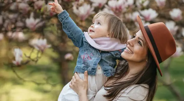 Uśmiechnięta mama w kapeluszu trzyma dziecko podczas sesji wiosennej dzieci. Maluch wskazuje palcem na kwitnącą magnolię.