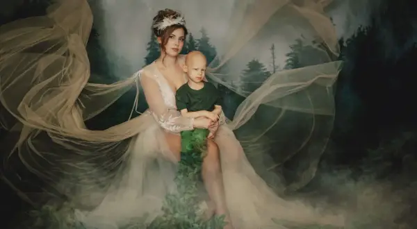 Ewa Miketa, niczym leśna nimfa, tuli chłopca w objęciach. Mglisty las i zwiewne tkaniny tworzą magiczną scenerię.