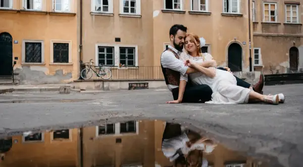 Kamil Włodarczyk i jego ukochana siedzą na ziemi, otoczeni starą kamienicą. Ich odbicie w kałuży dodaje magii tej chwili.