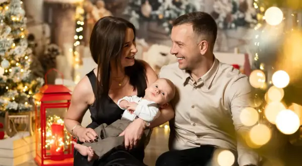 Rodzina podczas sesji bożonarodzeniowych. Mama i tata patrzą na siebie z uśmiechem, trzymając niemowlę. W tle choinki i lampki.