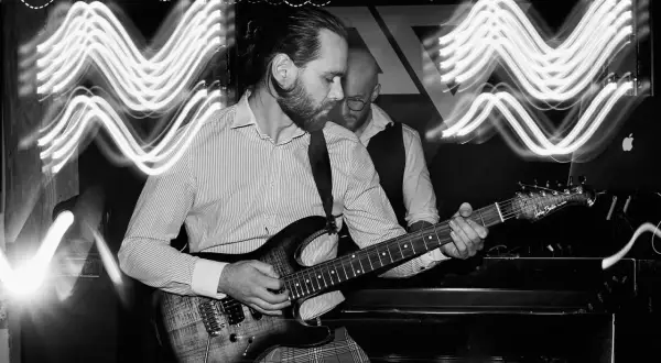Muzyk gra na gitarze elektrycznej, a za nim widać drugiego członka zespołu. Efekt falujących świateł sugeruje kompensację ekspozycji.
