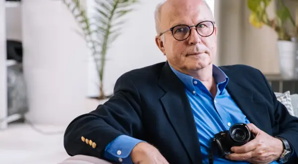 Tomasz Tomaszewski, w okularach i granatowej marynarce, trzyma aparat fotograficzny.