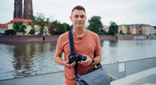 Mężczyzna z aparatem i torbą na ramieniu, gotowy do robienia zdjęć na dworze. W tle rzeka i zabytkowe budynki.