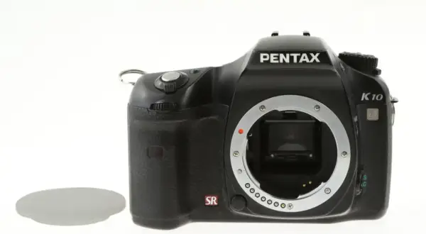 Czarny aparat Pentax K10 bez obiektywu, widoczna jego cmos matryca. Obok leży osłona.