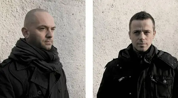 Szymon Rogiński w czarnym płaszczu i szalu, obok niego drugi mężczyzna w czarnej kurtce, obaj na tle chropowatej ściany.