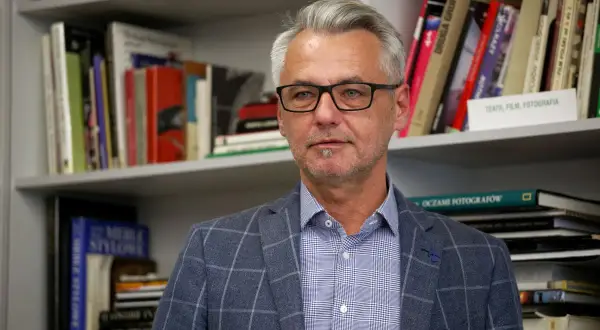 Andrzej Grygiel w okularach, w kratowaną marynarkę, na tle regałów z książkami o sztuce i fotografii.