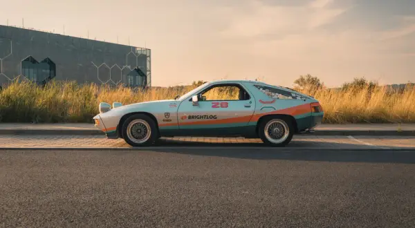 Klasyczna fotografia samochodowa: Porsche 928 w kolorach retro, z numerem 28 i logiem Brightlog, na tle nowoczesnego budynku i złotej trawy.