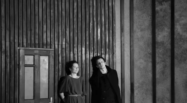Dwoje aktorów w czerni, w dramatycznej pozie, na tle drewnianej ściany i drzwi. Ta fotografia teatralna uchwyciła moment napięcia.