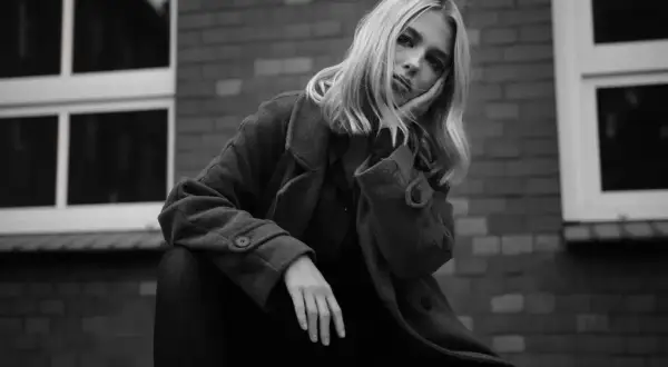 Blondynka w płaszczu opiera głowę na dłoni, zamyślona. Ten detal co to znaczy?