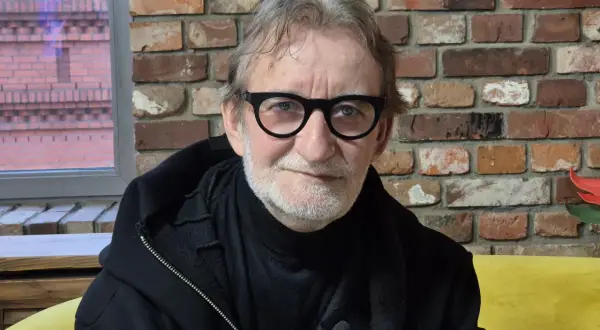 Maciej Mańkowski w okularach, z siwą brodą, w czarnej bluzie na tle ceglanej ściany.