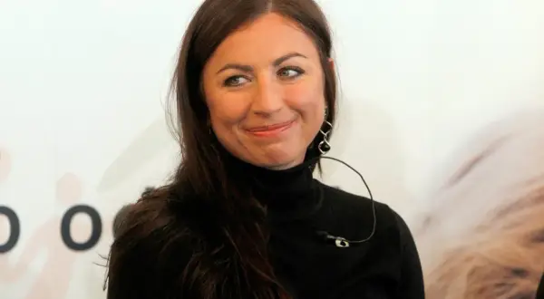 Uśmiechnięta Diana Kowalczyk w czarnym golfie, z charakterystycznymi kolczykami, patrzy w bok.