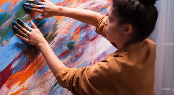 Ręce artystki pokryte niebieską i pomarańczową farbą tworzą kompozycję dynamiczną fotografię na abstrakcyjnym płótnie.
