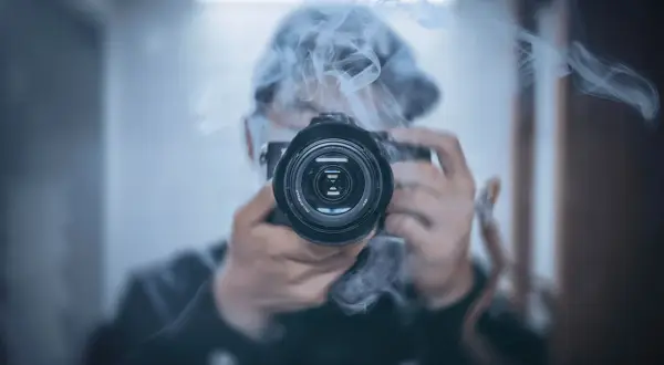 Osoba w kapturze śledzi i fotografuje gwiazdy przez obiektyw aparatu, otoczona mgiełką dymu.
