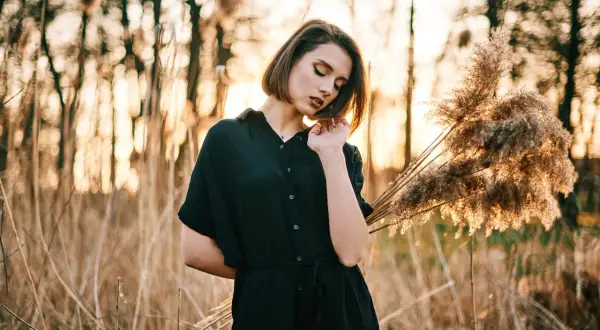 Młoda kobieta w czarnej sukience pozuje na tle zachodzącego słońca i trzcin. Idealna inspiracja na kurs fotografii portretowej.