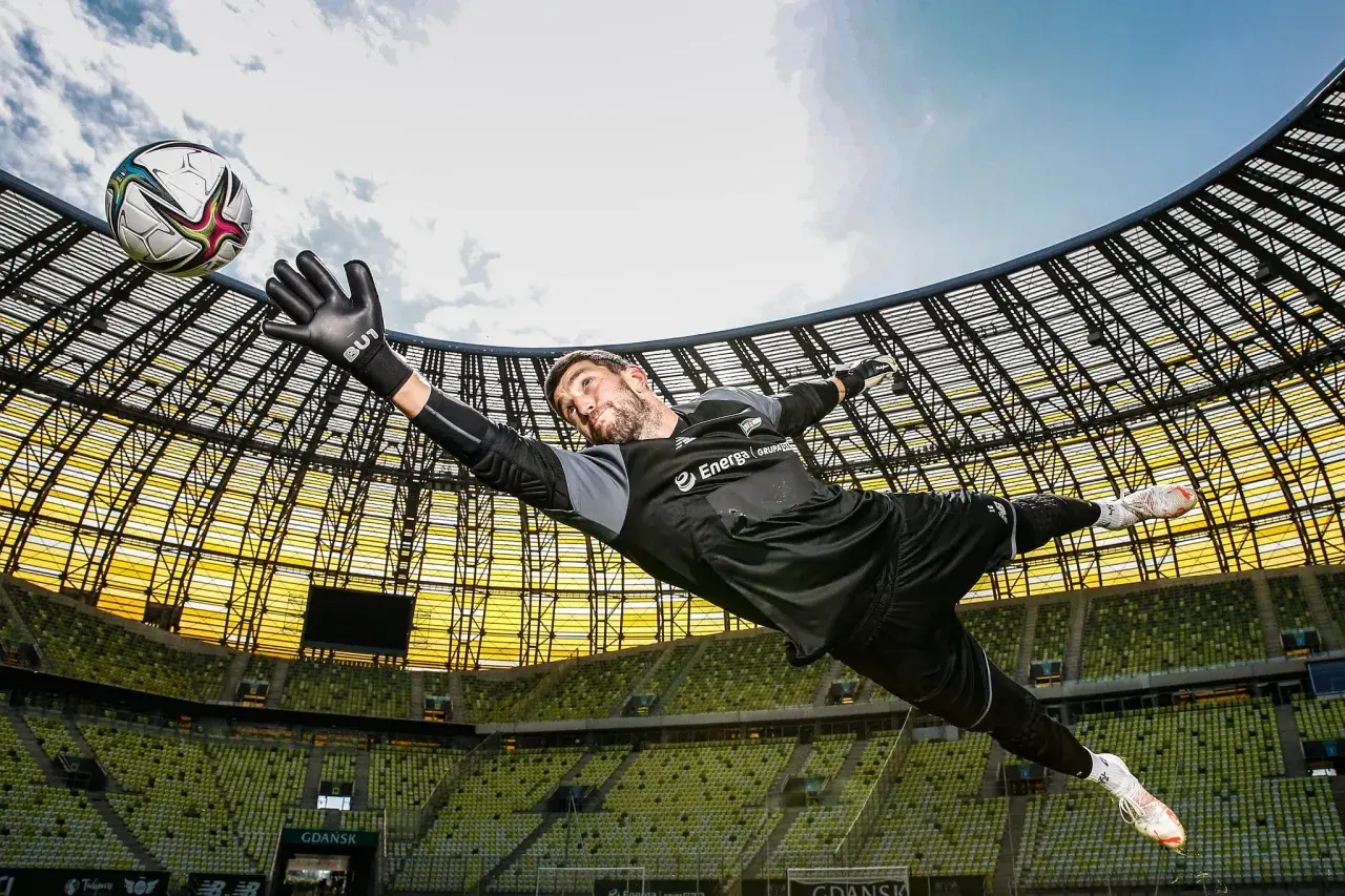 Bramkarz w locie łapie piłkę. Dynamiczna fotografia sportowa na tle stadionu.