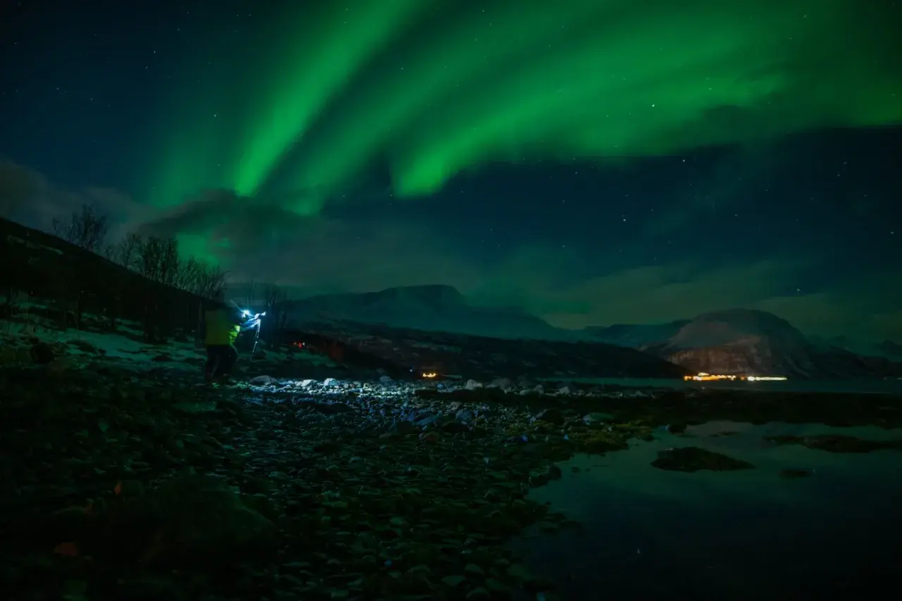 Nocne niebo rozświetla zorza polarna. Na pierwszym planie widać fotografa z lampą, który robi zdjecie z fleszem.