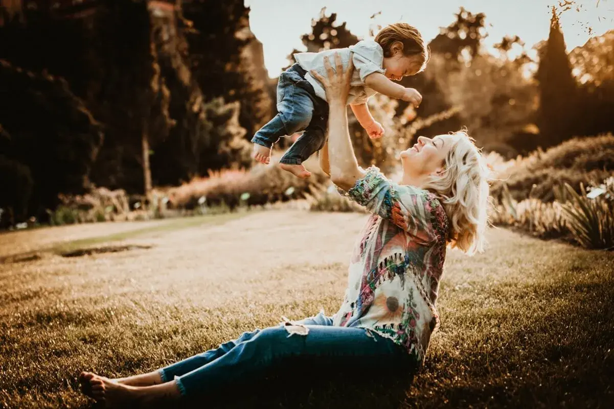 Mama rzuca dzieckiem w górę podczas śmiesznej sesji rodzinnej w parku. Słońce zachodzi, tworząc ciepłą, złotą poświatę.