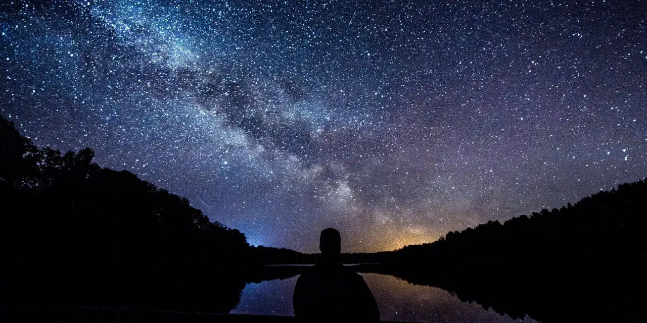 Sylwetka człowieka patrzy na Drogę Mleczną nad jeziorem. Idealny kadr, by pokazać, jak fotografować gwiazdy.