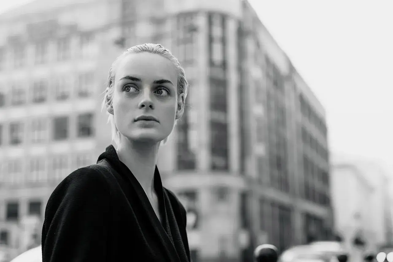 Monochromatyczna fotografia. Młoda kobieta o blond włosach patrzy w bok, na tle miejskiej architektury.