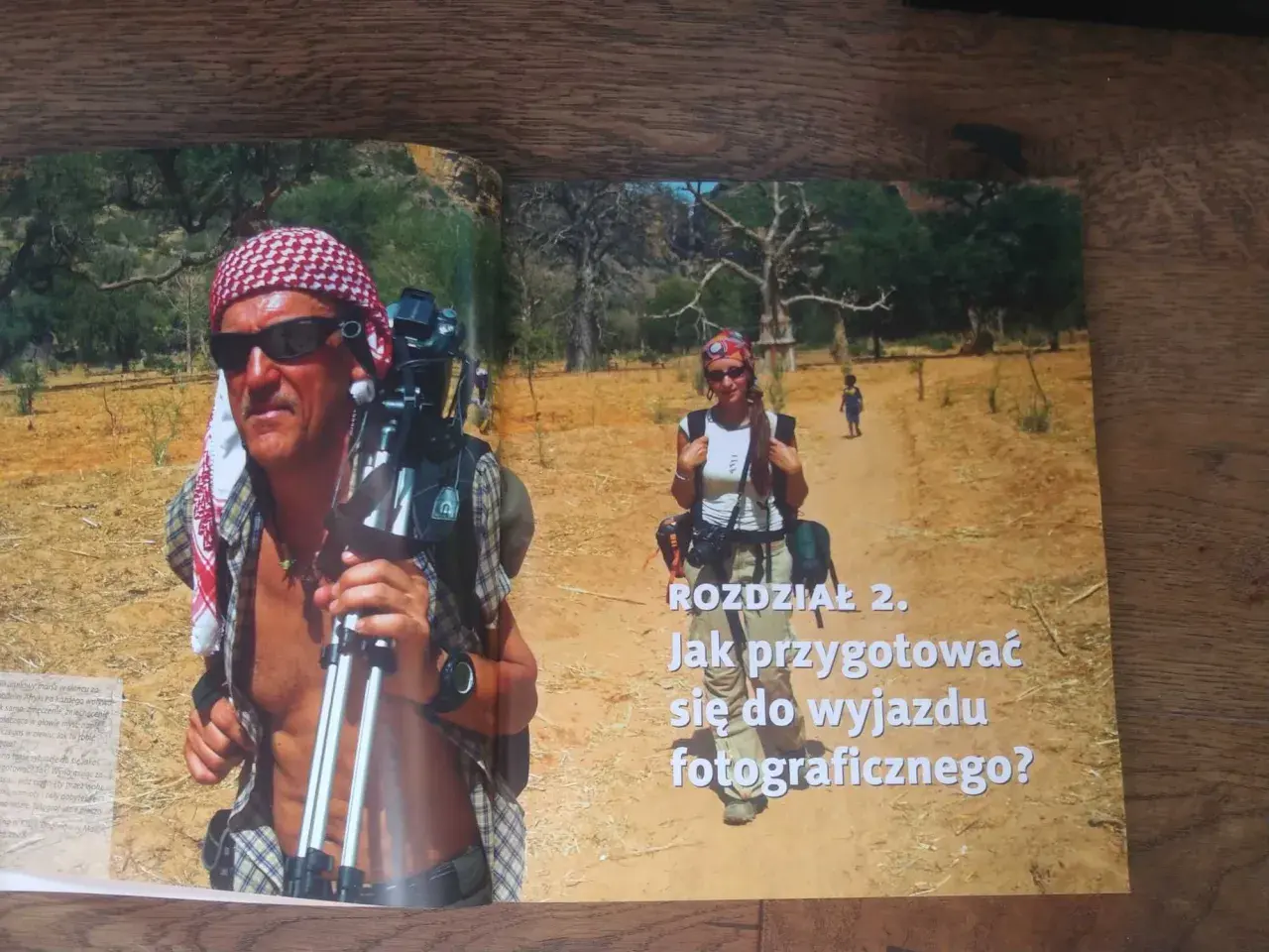 Piotr Trybalski z aparatem i statywem w gorącej Afryce. Obok niego kobieta i dziecko na pustynnej drodze.