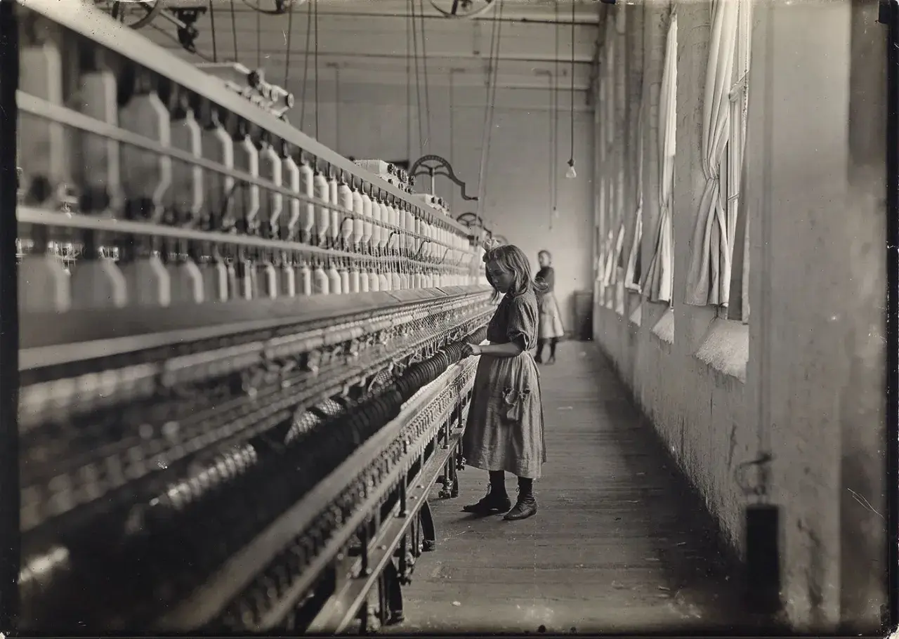Dziewczynka pracuje przy maszynie w fabryce, jak na zdjęciach Lewis Hine. W tle widać inną pracownicę.