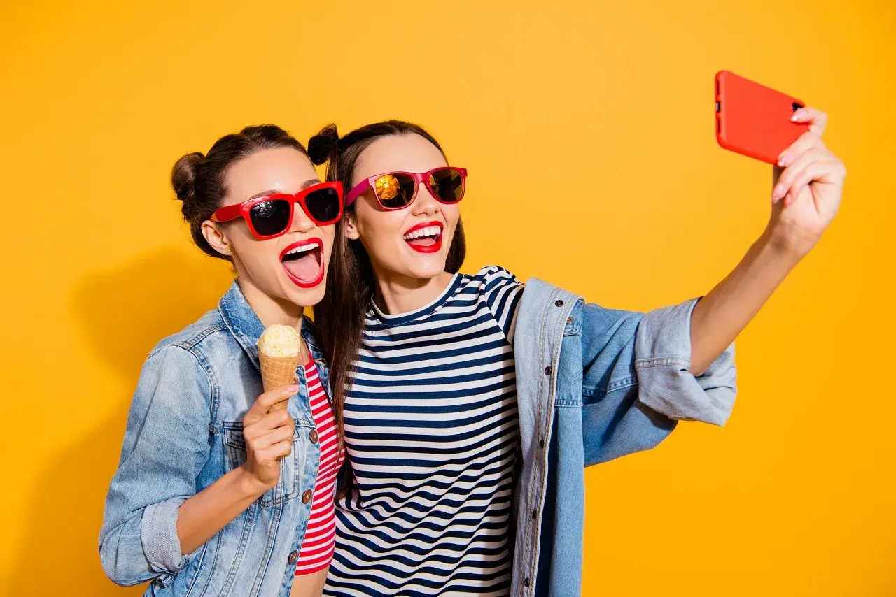 Dwie dziewczyny w okularach przeciwsłonecznych robią sobie selfie z lodami na patyku. Mają świetny pomysł na selfie!